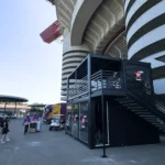 Struttura modulare Virgin Radio a due piani con pareti vetrate, scala di accesso e terrazza superiore, installata in prossimità delle tribune dello stadio per eventi musicali.