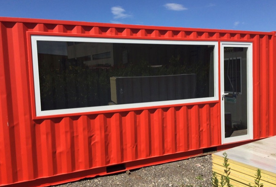 Container officina rosso con ampia finestra e porta vetrata, posizionato in un'area esterna.