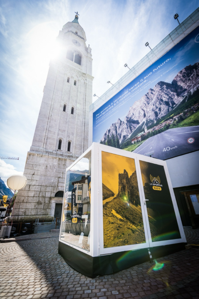 Showbox La Sportiva Radio posizionato accanto a un’iconica torre in pietra, con sole e montagna sullo sfondo, creando un contrasto tra modernità e tradizione.