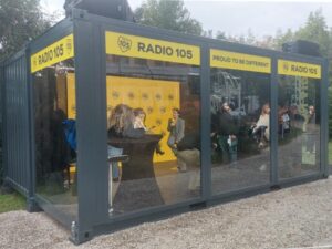 Container studio trasparente di Radio 105 allestito per Sanremo 2025, con persone all’interno che partecipano a un evento live.