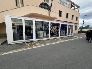 Esterno del showbox container per i 160 anni della Guardia Costiera a Sanremo, posizionato davanti alla Capitaneria di Porto, con ampie vetrate e grafica celebrativa.