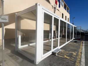 Prospettiva esterna del container vetrato allestito per il 160° anniversario della Guardia Costiera, con bandiere istituzionali e accesso facilitato per i visitatori.