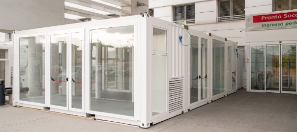 Struttura modulare in container bianco con pareti in vetro, installata all'esterno di un pronto soccorso per l'ampliamento delle strutture sanitarie.