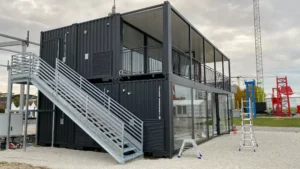 Vista laterale della struttura containerizzata a due piani progettata da Phoenix International per Jekko, con scala esterna metallica e vetrate panoramiche, durante la fase di allestimento a Bauma 2025.