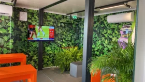 Zona green dello stand Jekko con pareti rivestite in fogliame artificiale, arredi arancioni e schermo TV, realizzata da Phoenix International per Bauma 2025.