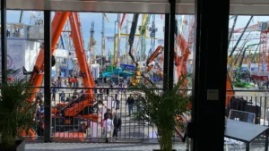 Vista dall’interno dello stand Jekko verso il piazzale esterno della fiera Bauma, con gru e macchinari edili esposti e visitatori in movimento.