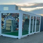 Struttura containerizzata Eat Natural con rivestimento bianco, pareti vetrate e pavimento in prato sintetico, fotografata al tramonto. Una soluzione espositiva modulare e scenografica per eventi promozionali e marketing itinerante.