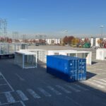 Container bianchi e blu posizionati nell’area esterna di EICMA 2025 durante la fase di allestimento dell’area Your Urban Mobility, con camion e moduli showbox pronti per l’installazione.