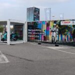 Container espositivo allestito per Yamaha nell’area Y.U.M. di EICMA 2025, con scooter elettrici esposti in un modulo vetrato e container brandizzato con grafica promozionale.