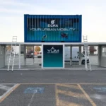 Struttura modulare a due livelli composta da container sovrapposti per l’area Y.U.M. di EICMA 2025, in fase di montaggio con scale appoggiate ai moduli e grafica ufficiale Your Urban Mobility in evidenza.