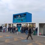 Ingresso dell’area Y.U.M. di EICMA 2025 con container espositivi e modulo sovrapposto brandizzato “Your Urban Mobility”, circondato da visitatori durante l’evento.