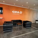 Area lounge interna della struttura CIFA con parete arancione brandizzata, divani neri e schermo.