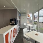 Area hospitality interna con bancone, tavoli, sedute e ampie vetrate affacciate sull’esterno.