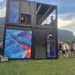 Vista laterale della struttura Vigorsol con container sovrapposti, terrazza illuminata e rivestimenti grafici blu e rossi.